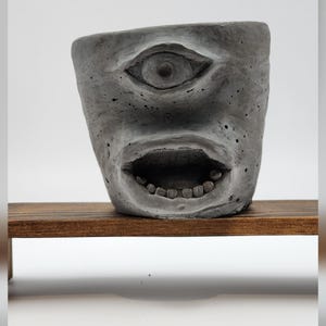 May include: A gray concrete planter shaped like a cyclops head with a single eye centered above an open mouth featuring visible teeth. The surface of the planter has a porous, textured finish with small indentations throughout the material.
