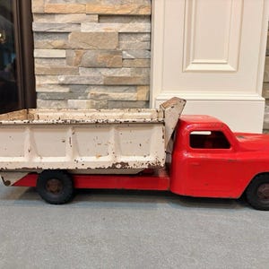 May include: A vintage metal toy dump truck featuring a bright red cab and a white dump bed with visible paint wear and rust. The truck has black wheels and an open cab design, showcasing a classic construction vehicle aesthetic with signs of age.