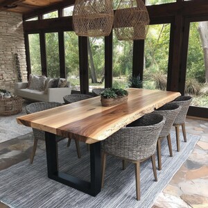 Puede incluir: Una mesa de comedor rectangular con una superficie de madera natural de borde irregular y patas de metal negro en forma de U. La mesa est&aacute; rodeada por seis sillas de mimbre trenzado gris con patas de madera y una cesta con plantas verdes.