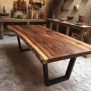 Puede incluir: Una mesa de comedor rectangular con un tablero de madera maciza de borde natural, con vetas y nudos visibles. La mesa se apoya sobre dos patas de metal negro en forma de U, que proporcionan una base estable para la gruesa superficie de madera.