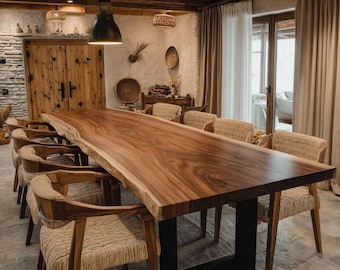 Puede incluir: Una mesa de comedor larga con un tablero de madera maciza de borde natural y una base de metal negro en forma de U. La mesa está rodeada por varias sillas de madera con asientos y respaldos trenzados en una habitación con paredes de piedra.