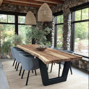 Puede incluir: Una mesa de comedor rectangular con una superficie de madera natural de borde vivo y una base de metal negro en forma de U. Seis sillas grises tapizadas con patas negras est&aacute;n dispuestas alrededor. Dos l&aacute;mparas colgantes tejidas.