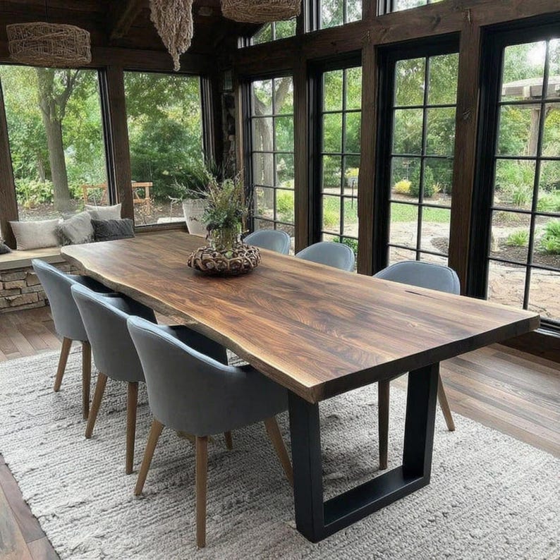 Puede incluir: Una mesa de comedor rectangular grande con una superficie de madera natural de borde irregular y dos patas de metal negro en forma de U. La mesa est&aacute; rodeada por seis sillas tapizadas en gris con patas de madera sobre una alfombra gris claro.
