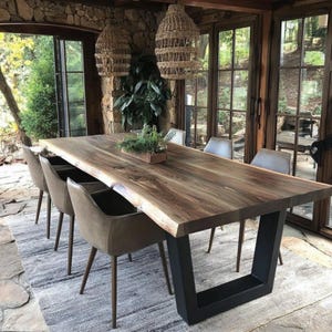 Puede incluir: Una gran mesa de comedor rectangular con una superficie de madera natural de borde vivo y patas de metal negro en forma de U. La mesa est&aacute; rodeada por cuatro sillas tapizadas en gris con patas c&oacute;nicas. Dos l&aacute;mparas colgantes de mimbre cuelgan.