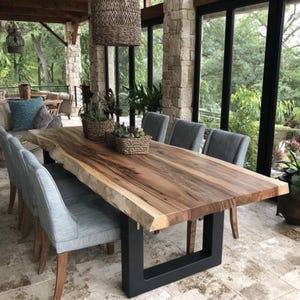 Puede incluir: Una mesa de comedor grande con una superficie de madera natural de borde irregular y patas de metal negro en forma de U. La mesa est&aacute; rodeada por seis sillas grises tapizadas. Dos cestas tejidas con plantas verdes descansan sobre la mesa.