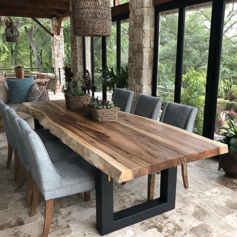 Puede incluir: Una mesa de comedor grande con una superficie de madera natural de borde irregular y patas de metal negro en forma de U. La mesa est&aacute; rodeada por seis sillas grises tapizadas. Dos cestas tejidas con plantas verdes descansan sobre la mesa.