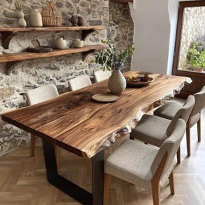 Könnte beinhalten: Ein Esstisch aus Holz mit Baumkante, natürlicher Maserung und schwarzen Metallbeinen, dazu sechs gepolsterte Stühle mit hellgrauen Stoffbezügen und Holzrahmen, aufgestellt in einem Raum mit Steinwänden und Holzregalen.