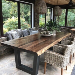 Puede incluir: Una gran mesa de comedor rectangular con una superficie de madera natural de borde irregular y una base de metal oscuro en forma de U. La mesa est&aacute; rodeada de sillones de mimbre trenzado y un banco con cojines grises estampados.
