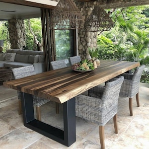 Puede incluir: Una mesa de comedor rectangular con una superficie de madera natural de borde vivo con vetas de color marr&oacute;n oscuro y una base robusta de metal negro en forma de U. La mesa est&aacute; rodeada por cuatro sillones de mimbre trenzado gris.