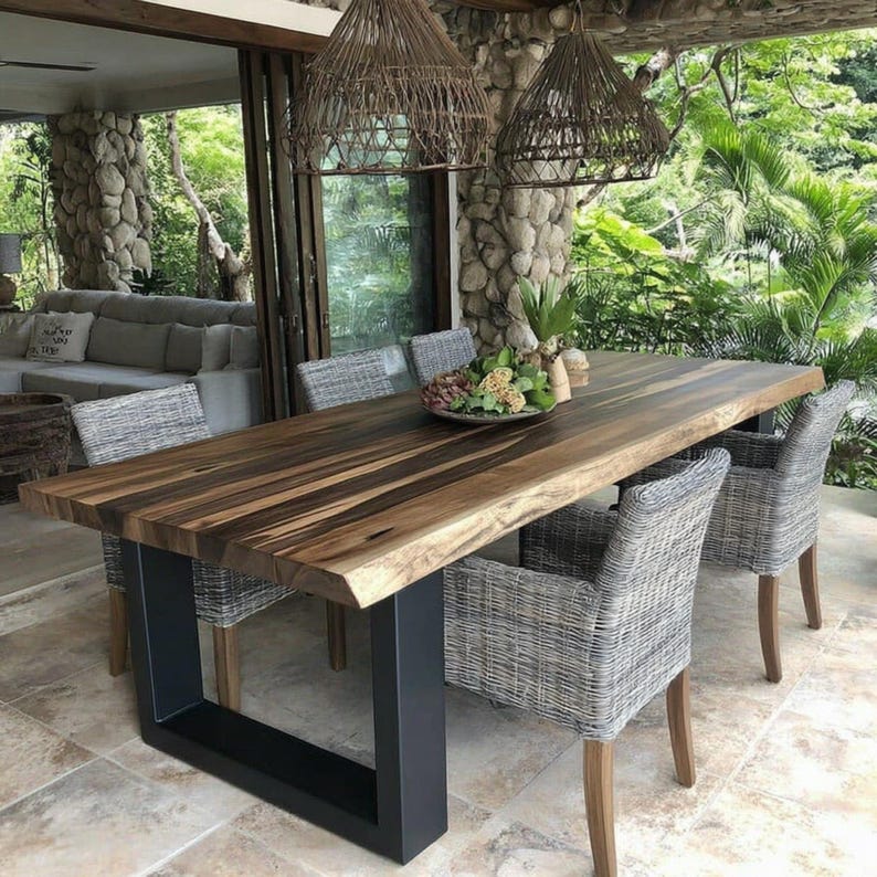 Puede incluir: Una mesa de comedor rectangular con una superficie de madera natural de borde vivo con vetas de color marr&oacute;n oscuro y una base robusta de metal negro en forma de U. La mesa est&aacute; rodeada por cuatro sillones de mimbre trenzado gris.