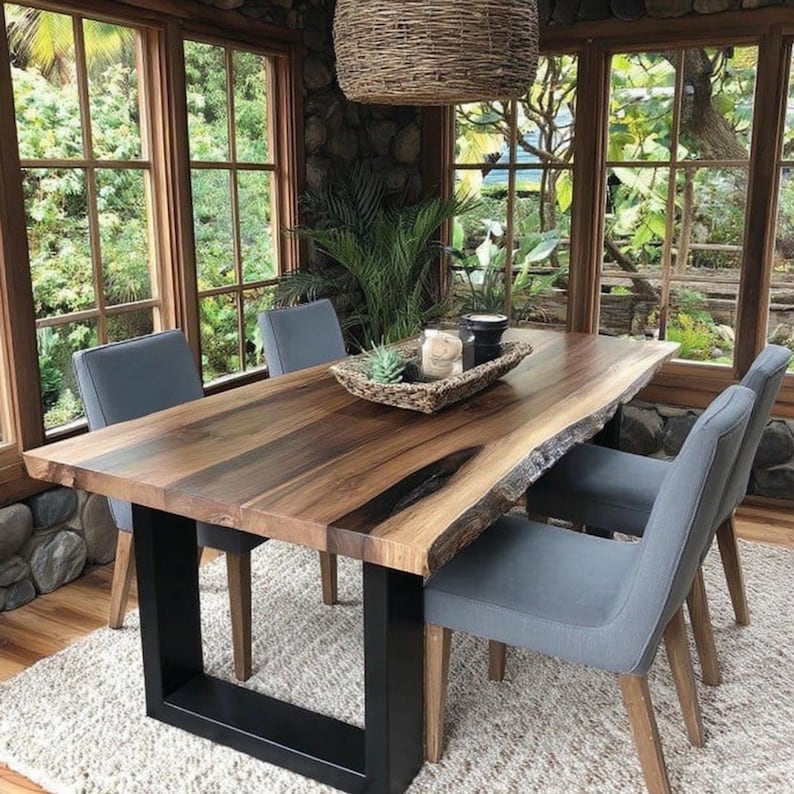 Puede incluir: Una mesa de comedor rectangular con un tablero de madera de borde natural con vetas de color marr&oacute;n oscuro, sostenida por dos patas de metal negro en forma de U. Cuatro sillas tapizadas en gris con patas de madera rodean la mesa.
