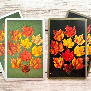May include: Playing cards with a design of autumn leaves in red, orange, and yellow. The cards have a green and black background, with a white border. The leaves are arranged in a decorative pattern.