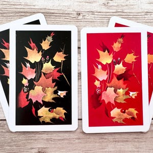 May include: Playing cards with white borders display a design of autumn leaves in red, orange, and yellow hues. One set of cards has a black background, and the other has a red background. The cards are arranged on a wooden surface.