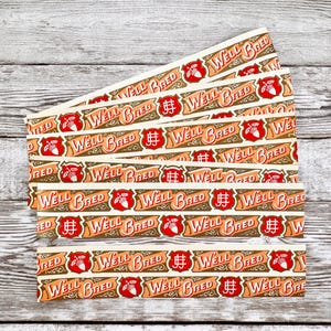 May include: Several paper strips featuring the text "Well Bred" in a vintage style. The design includes a red shield with an acorn and wheat, set against a brown and cream background. The strips are arranged on a weathered wooden surface.
