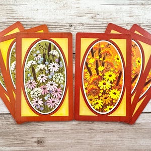 May include: A set of vintage playing cards with floral designs. Each card has a yellow background with an oval floral illustration in the centre, framed by a red border. The floral designs feature white, pink, and yellow flowers.