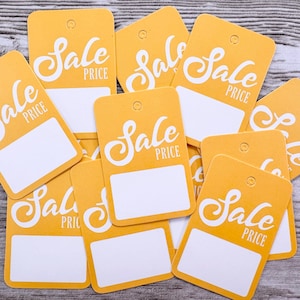 May include: A pile of yellow price tags, each with the words "Sale Price" in white script. Each tag has a white rectangular space for writing. The tags are scattered on a light wood-grain surface.