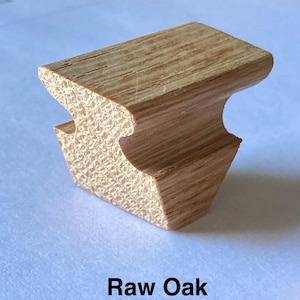 May include: A small, unfinished piece of light brown oak wood with a distinct grain pattern. The object features a wide, flat top surface that tapers inward to a narrower base with curved side indentations. The text Raw Oak is printed below.