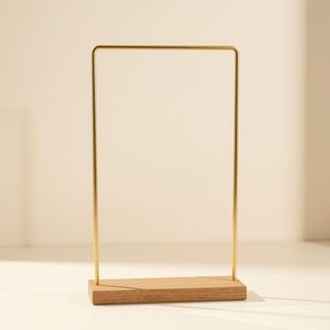 May include: A vertical gold-toned metal frame with rounded top corners, securely mounted into a rectangular light-colored wooden base. The structure features a clean, open design with a smooth finish on both the metal and the wood components.