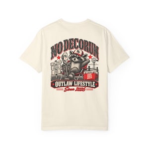 May include: An off-white short-sleeve t-shirt featuring a graphic on the back of a raccoon wearing sunglasses and a cap, sitting in a folding chair with a drink. The text reads NO DECORUM, BAD IDEAS, BETTER COMPANY, SAME STORY, OUTLAW LIFESTYLE, SINCE 2026, and GOOD TIMES AHEAD.