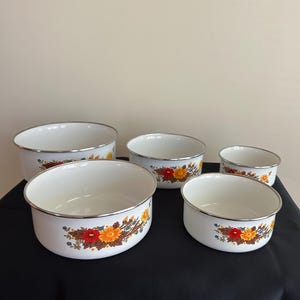 Peut inclure: Un ensemble de cinq bols en émail blanc avec des bords argentés, ornés d'un motif floral composé de fleurs rouges, orange et jaunes avec des feuilles brunes. Les bols sont disposés en groupe, variant de taille, avec une finition lisse.