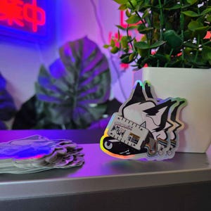 May include: A holographic sticker featuring a stylized black and white animal head wearing a white rectangular device with the text gutworx, MCM, and a warning symbol, connected by a rainbow-colored cable, with a stack of identical stickers nearby.