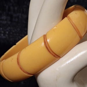 Op de afbeelding: Een gladde, lichtgele armband met een gesegmenteerd ontwerp. De armband heeft afgeronde, gebogen secties gescheiden door dunne, verzonken bruine lijnen die een herhalend patroon vormen rond de gehele omtrek van de cirkelvormige band.