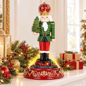 May include: A decorative nutcracker figurine wearing a red and gold crown, a green jacket, and red trousers. The figure holds a small green Christmas tree and stands on a circular red base featuring a miniature train and two golden spotlights.