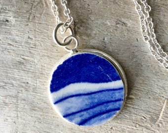 May include: A silver chain necklace with a round pendant featuring a blue and white ceramic design. The pendant has a white background with blue lines and a blue border.
