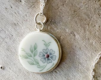 May include: A silver chain necklace with a round pendant featuring a white porcelain background with a green floral design and a purple center.