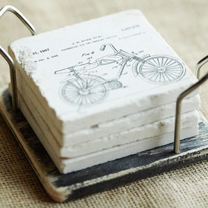 Puede incluir: Conjunto de cuatro posavasos de cer&aacute;mica blanca con un dibujo de patente de motocicleta vintage en blanco y negro. Los posavasos est&aacute;n en un soporte de madera negro con un asa de metal.