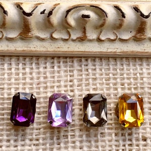 Puede incluir: Cuatro gemas de vidrio de forma rectangular en diferentes colores. Las gemas son de color púrpura, azul claro, gris y amarillo. Las gemas están colocadas sobre un fondo de arpillera.