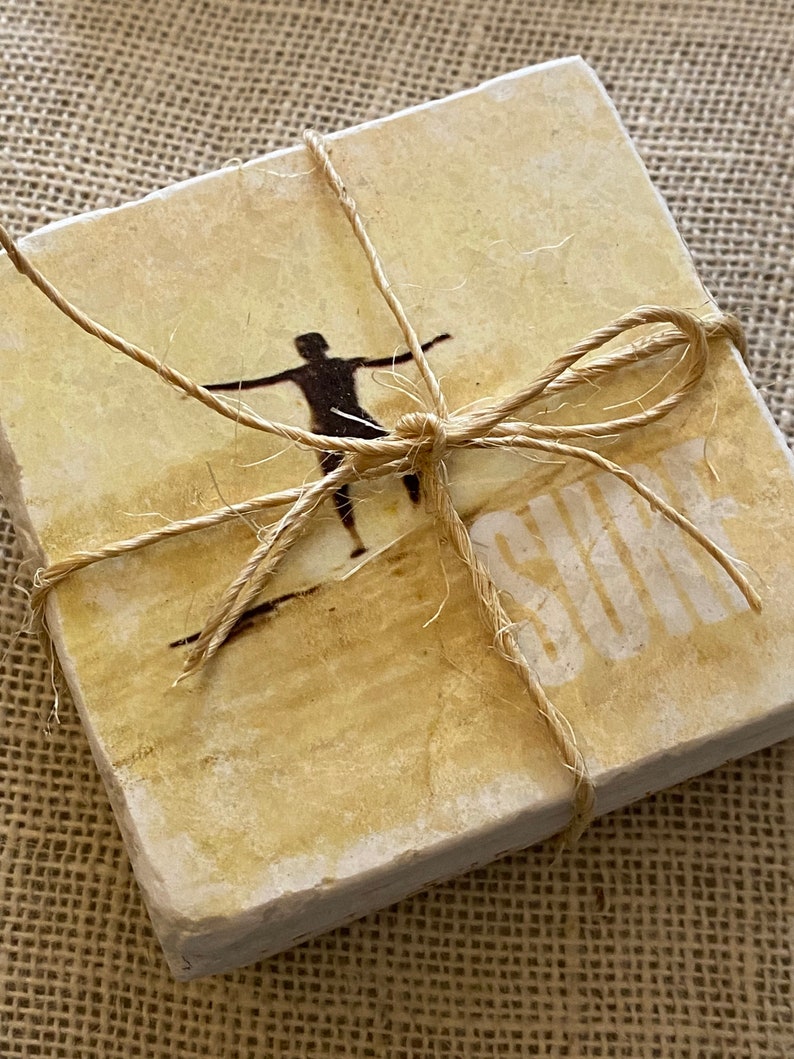 Pode incluir: Conjunto de quatro  coasters bege com uma silhueta de uma pessoa surfando e a palavra "SUN" impressa neles. Os coasters est&atilde;o amarrados com barbante.
