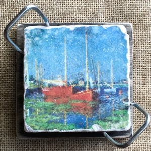 May include: A square coaster with a painted image of red sailboats on a blue body of water. The coaster is made of natural stone and has a rustic, handmade look.