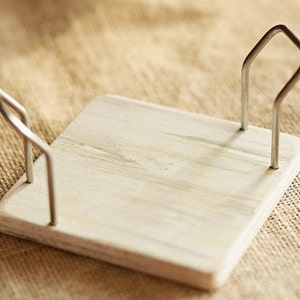 K&ouml;nnte beinhalten: Ein wei&szlig;er Serviettenhalter aus Holz mit zwei Metallschlaufen an jeder Seite. Der Halter steht auf einer braunen Jutestoffoberfl&auml;che.