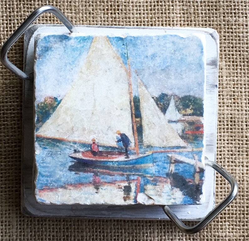 May include: A white wooden coaster with a painted image of a sailboat on a body of water. The coaster has a metal handle on each side.