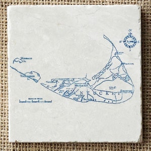 May include: A white ceramic coaster with a blue and white map of Nantucket Island, Massachusetts. The map includes the names of towns and landmarks, such as Madaket, Sconset, and Nantucket Harbor. A compass rose is in the upper right corner.