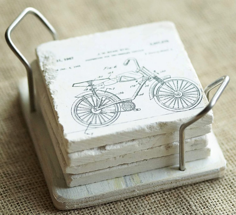 Puede incluir: Conjunto de posavasos de cer&aacute;mica blanca con un dibujo de patente de bicicleta vintage. Los posavasos est&aacute;n apilados y unidos por un soporte de metal.