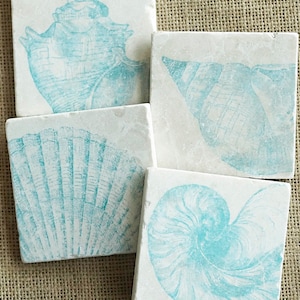 May include: Set of four white square coasters with blue seashell designs. Each coaster features a different seashell, including a conch shell, a scallop shell, and a nautilus shell.