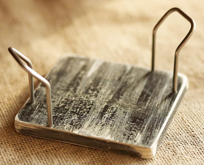 K&ouml;nnte beinhalten: Ein rustikaler Holz-Serviettenhalter mit einer abgenutzten schwarzen und wei&szlig;en Oberfl&auml;che. Der Halter hat zwei Metallschlaufen oben, um Servietten zu halten.