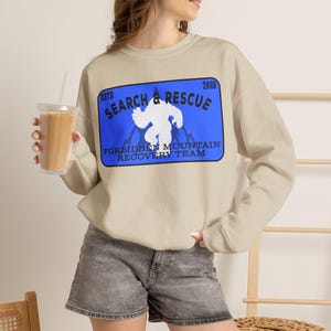 May include: A beige crewneck sweatshirt featuring a rectangular blue graphic on the chest. The graphic displays a white silhouette of a creature, the text "ESTD 2006", "SEARCH & RESCUE", and "FORBIDDEN MOUNTAIN RECOVERY TEAM" in black lettering.