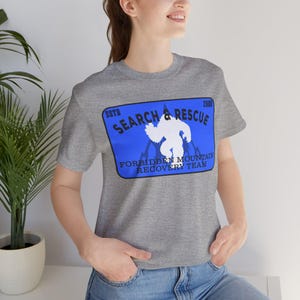 May include: A heather gray short-sleeve t-shirt featuring a blue rectangular graphic on the chest. The graphic displays a white silhouette of a creature, the text "SEARCH & RESCUE", "ESTD 2006", and "FORBIDDEN MOUNTAIN RECOVERY TEAM" in black.