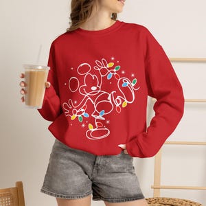 May include: A bright red long-sleeve sweatshirt featuring a white line-art graphic of Mickey Mouse tangled in colorful Christmas lights. The design includes small starburst accents and multi-colored light bulbs in red, blue, green, and yellow.