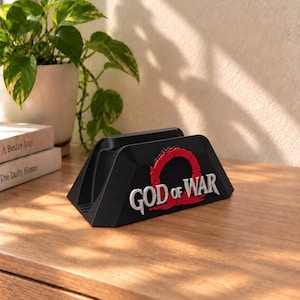 May include: A black controller stand featuring the God of War logo in raised white and red lettering on the front. The stand has a trapezoidal shape with a central slot designed to hold a gaming controller securely in a horizontal position.