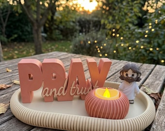 May include: A beige oval tray holds a coral-colored 3D sign with the text PRAY and trust, a small coral-colored ribbed candle holder with a yellow LED flame, and a small figurine of a person with brown hair and a white robe.
