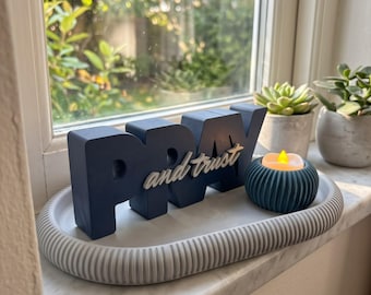 May include: A navy blue block letter sign reading PRAY with the white script text and trust attached to the front. The sign sits on a light gray oval ribbed tray next to a small dark teal ribbed candle holder with a glowing yellow flame.
