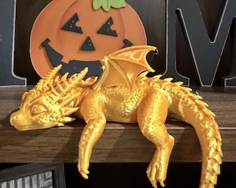 May include: A golden, articulated dragon figurine with textured scales and wings, resting on a ledge. The dragon has a detailed head with horns and a long, segmented tail that hangs down. Behind the dragon is a flat, orange pumpkin cutout.