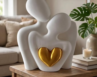 May include: A smooth white ceramic sculpture depicting two abstract figures embracing, with a prominent metallic gold heart positioned at the center of the figures. The sculpture features rounded, featureless forms and a glossy finish.