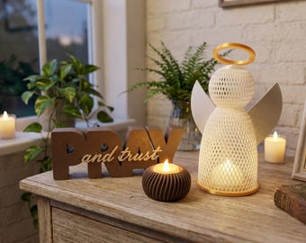 May include: A white 3D-printed angel figurine with a gold halo, a brown block sign reading PRAY and trust, and a small dark brown ribbed candle holder. The angel and candle holder contain glowing LED lights, creating a warm illumination.
