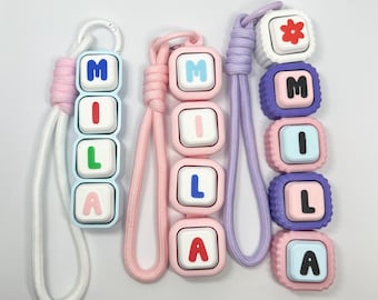 May include: Three keychains featuring square, button-like blocks arranged vertically. Each keychain spells the name MILA with colourful letters. The keychains have white, pink, and purple braided wrist straps attached to plastic rings.