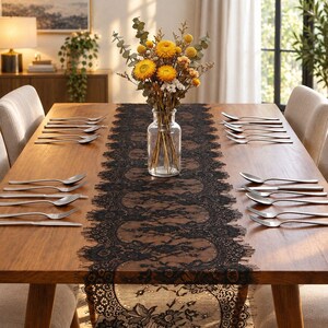 May include: A black lace table runner with a floral pattern and scalloped edges centered on a wooden dining table. The table is set with silver forks, knives, and spoons, and features a clear glass vase filled with yellow dried flowers.